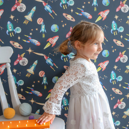 Child in a white dress standing against a wall with space-themed, rocket wallpaper
