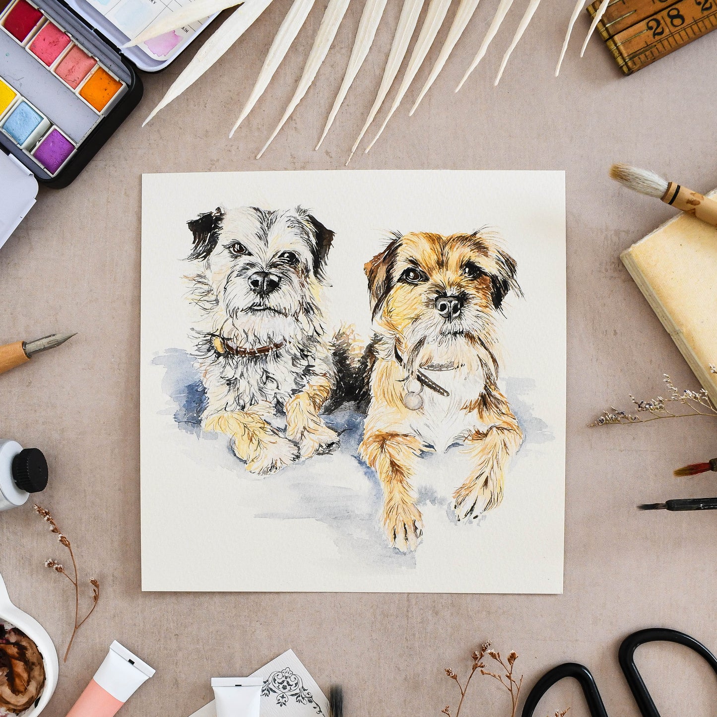 hand-painted portrait of two border terriers on a watercolour background with art supplies around.