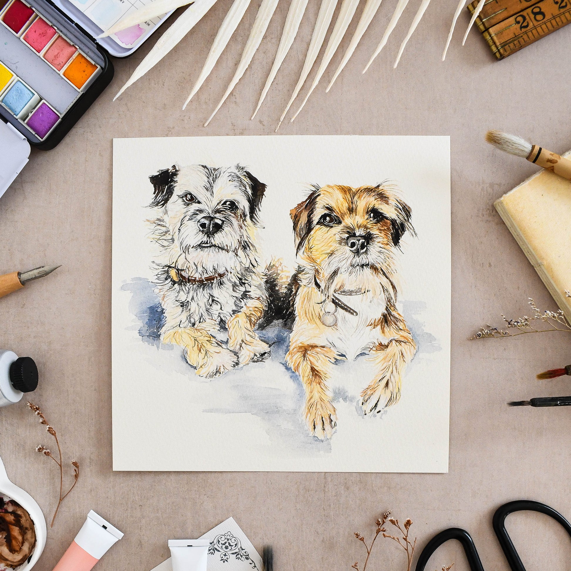 hand-painted portrait of two border terriers on a watercolour background with art supplies around.
