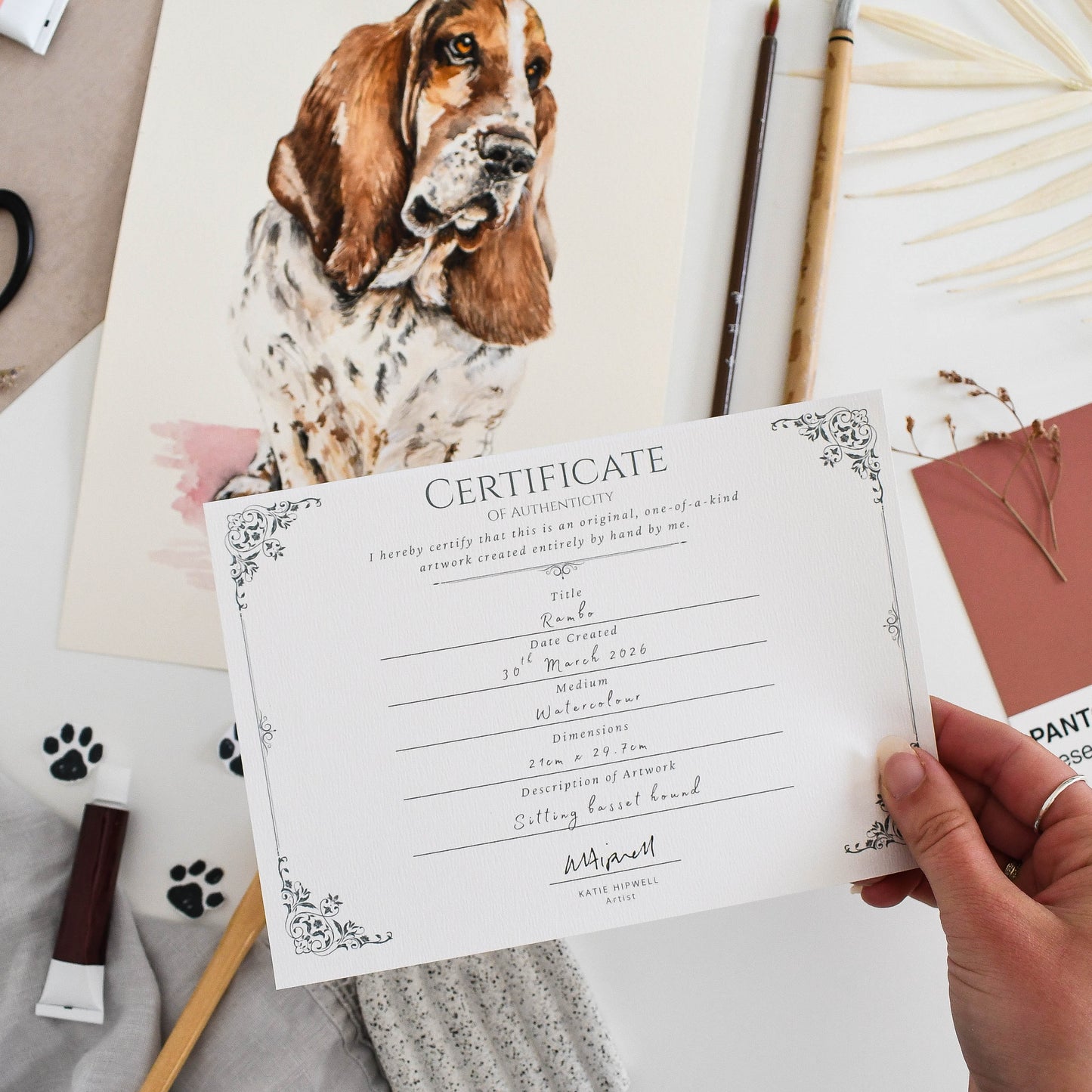Hand holding a certificate of authenticity with a basset hound dog painting on a table with art supplies