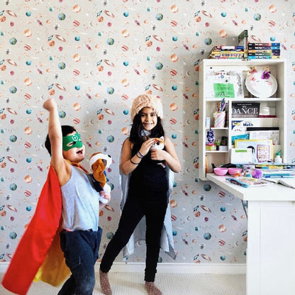 Children in superhero costumes in a room with colorful planets wallpaper and a desk.