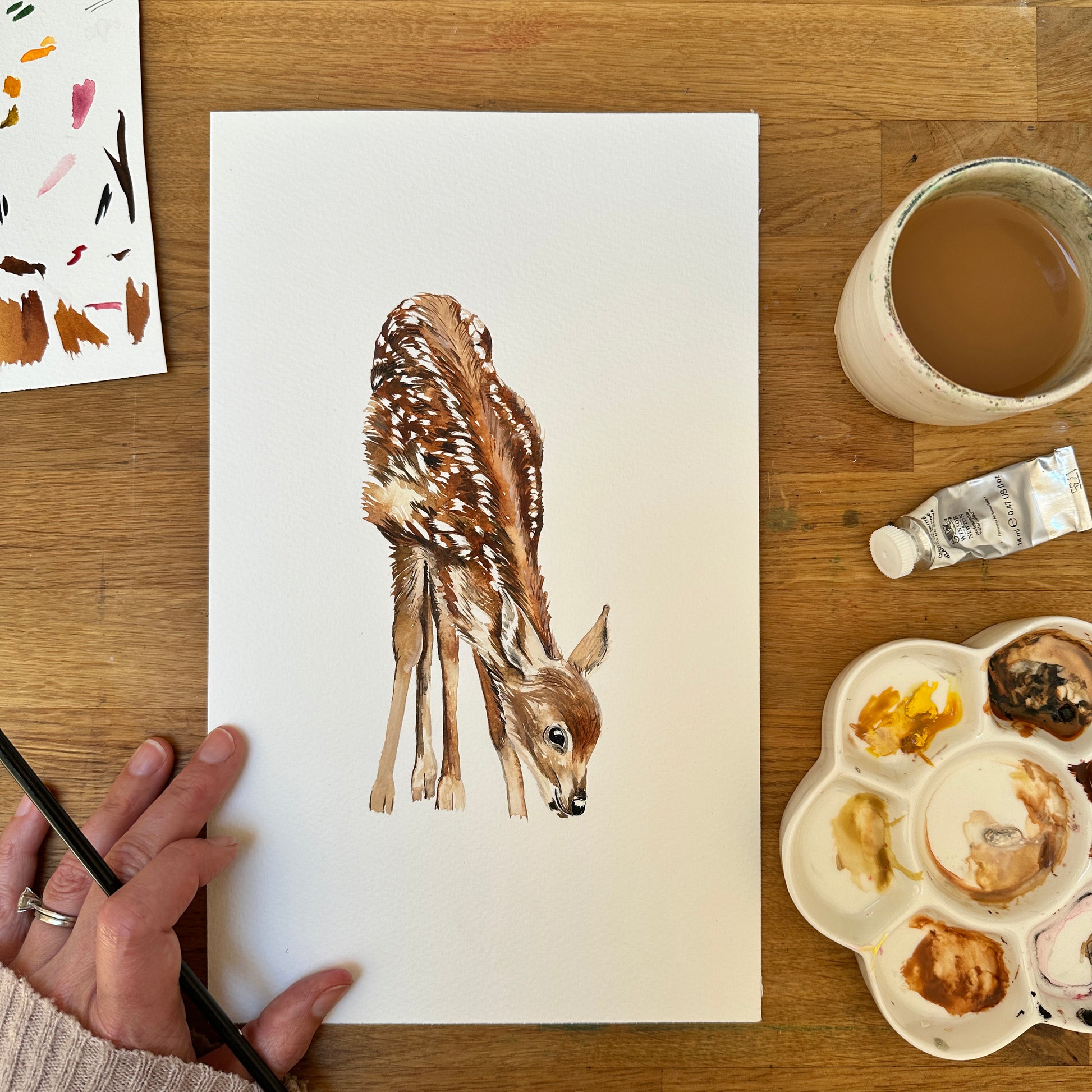 Watercolour paintings of a baby deer on paper with painting supplies on a wooden table
