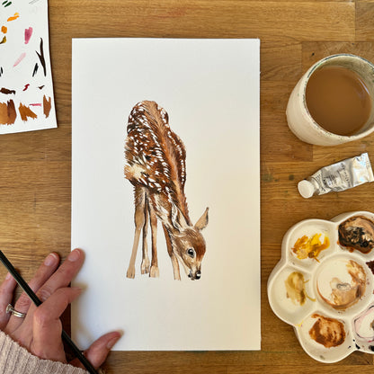 Watercolour paintings of a baby deer on paper with painting supplies on a wooden table